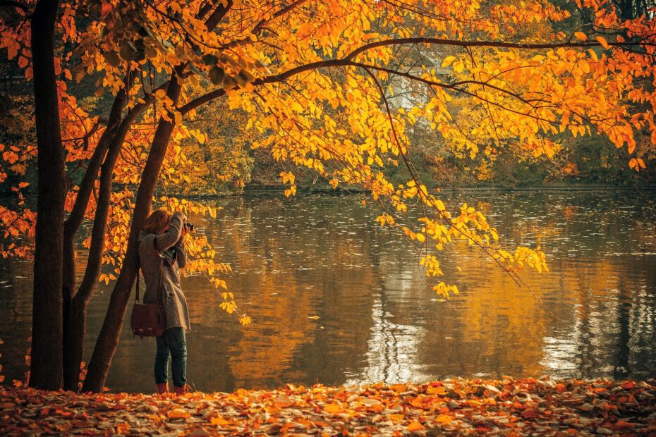 woman in brown coat standing near orange leafed tree and body of water during daytime